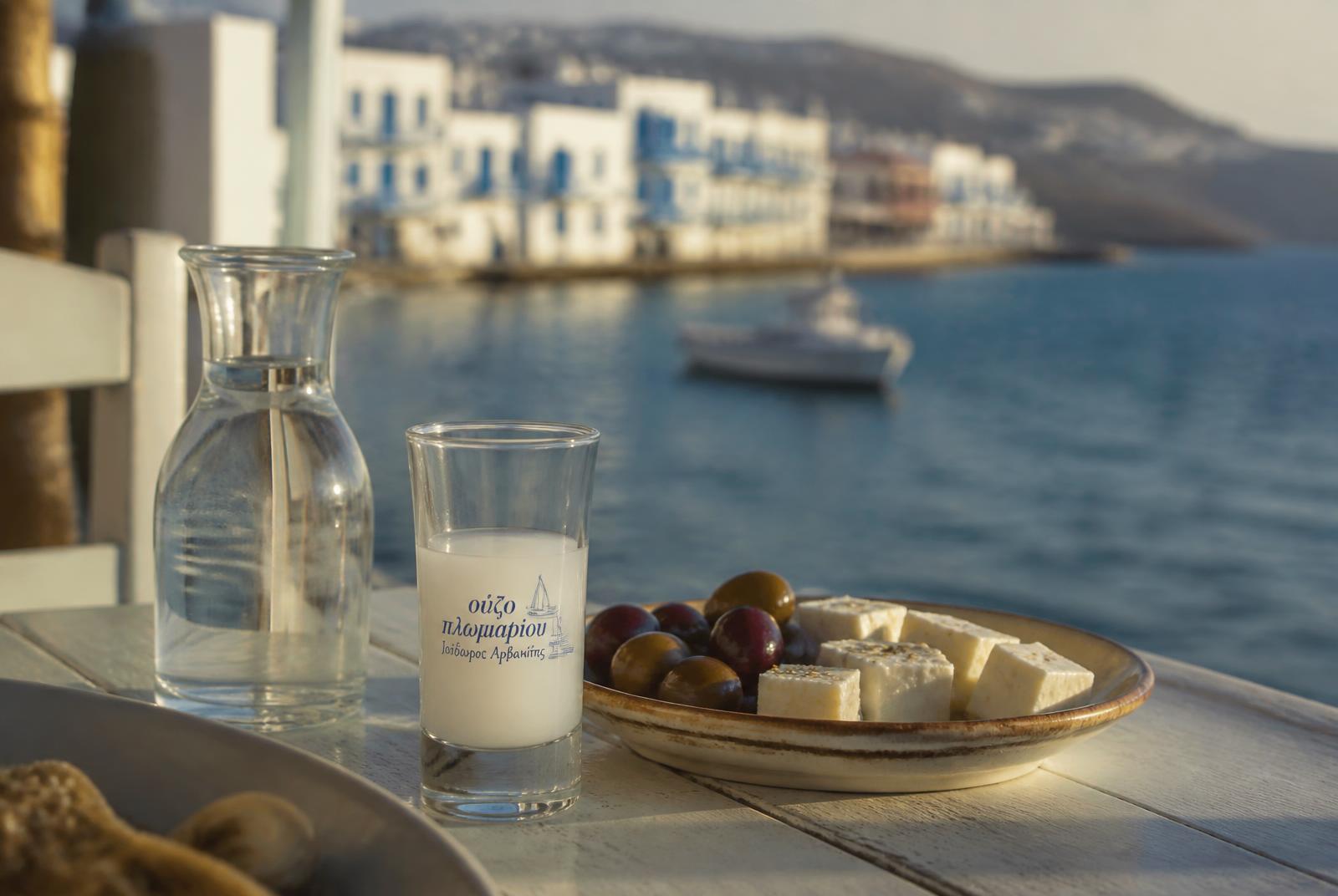 A glass of ouzo clouding to milky white next to a water carafe on a Greek taverna table.